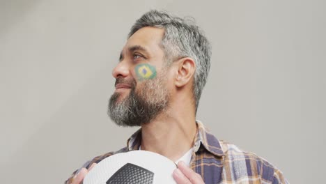 hombre biracial feliz con bandera de brasil en la mejilla sosteniendo un fútbol