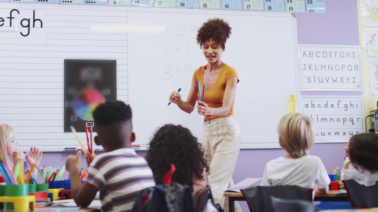 Female Teacher Standing At Whiteboard Teaching Maths Lesson To Elementary Pupils In School Classroom