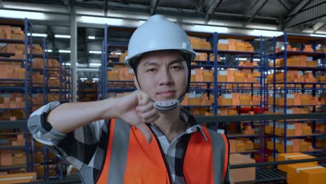 primer plano de un ingeniero masculino asiático con casco de seguridad de pie en el almacén con estantes llenos de productos de entrega. mostrando los pulgares hacia abajo y sacudiendo la cabeza en el almacenamiento
