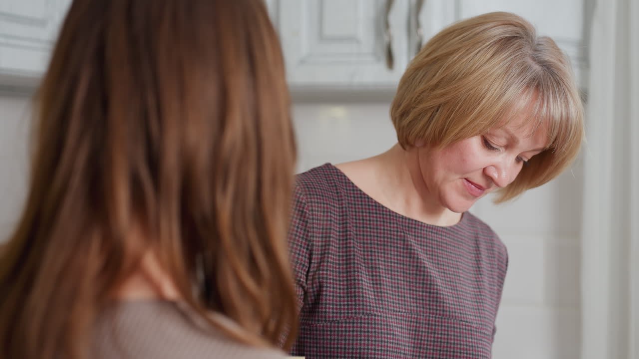 밝은 부에서 요리하는 이모를 쳐다보는 동안 책을 들고 있는 소녀의 근접 후면 뷰, 이모는 음식 준비에 집중하고 있으며, 소녀는 계속 읽기 전에 잠시 관찰합니다.
