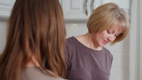 close-up rear view of girl holding book while glancing at aunt cooking in bright kitchen, aunt is focused on food preparation, while girl observes briefly before continuing reading