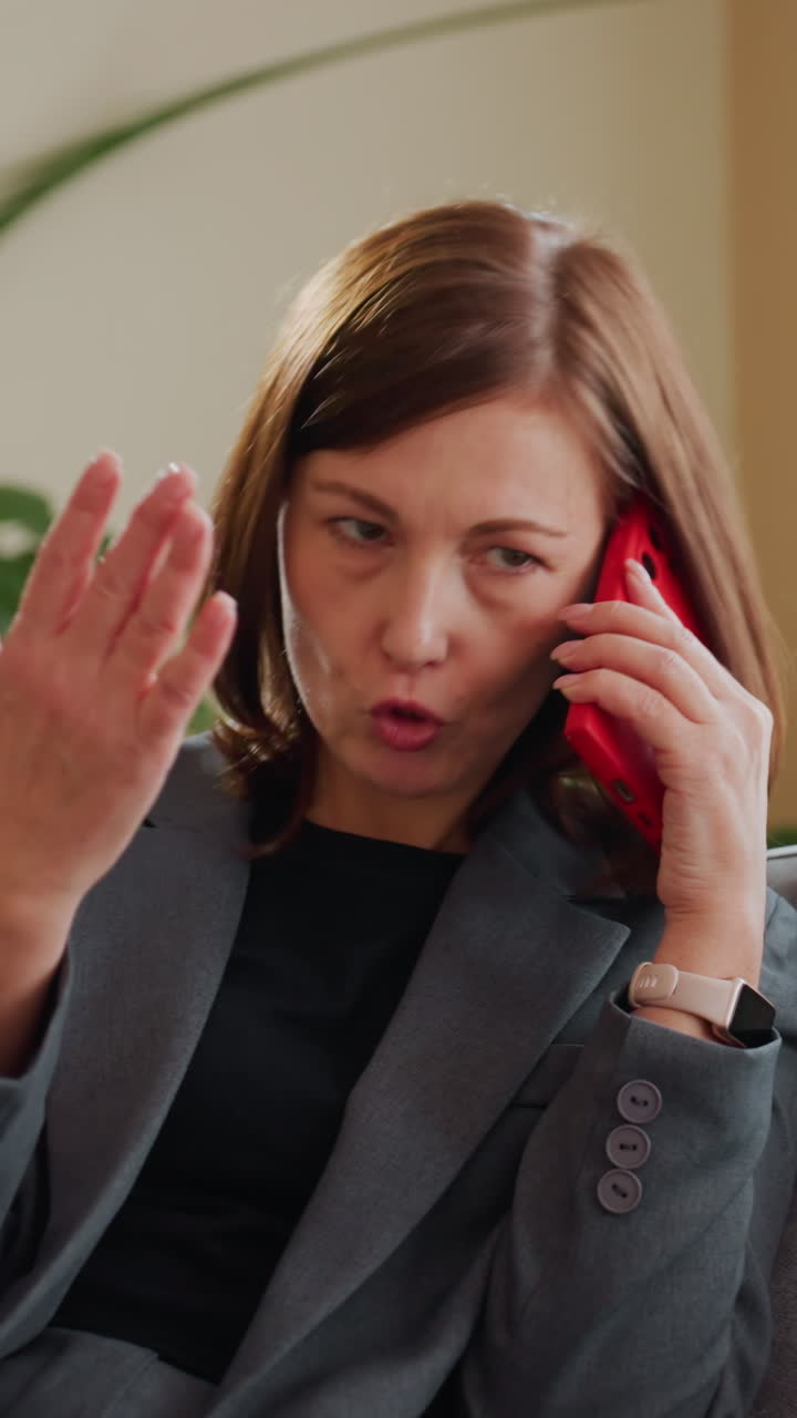 Frustrated businesswoman in gray suit talking on phone indoors gesturing emotionally while sitting on couch near green plants in modern office or home environment during daytime