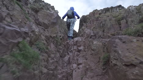 Dolly-shot-of-girl,-woman-climbing-rocky-mountain-cliff-at-dusk-in-Edinburgh,-Scotland-to-reach-Arthurs-seat