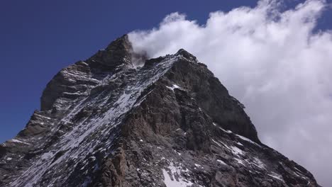 Vista-Aérea-Ascendente-Desde-Drones-Del-Matterhorn,-Famosa-Cima-Del-Monte-Cervino-Con-Cielo-Azul-Brillante-En-Un-Lado-Y-Nubes-Blancas-Suaves-En-El-Otro