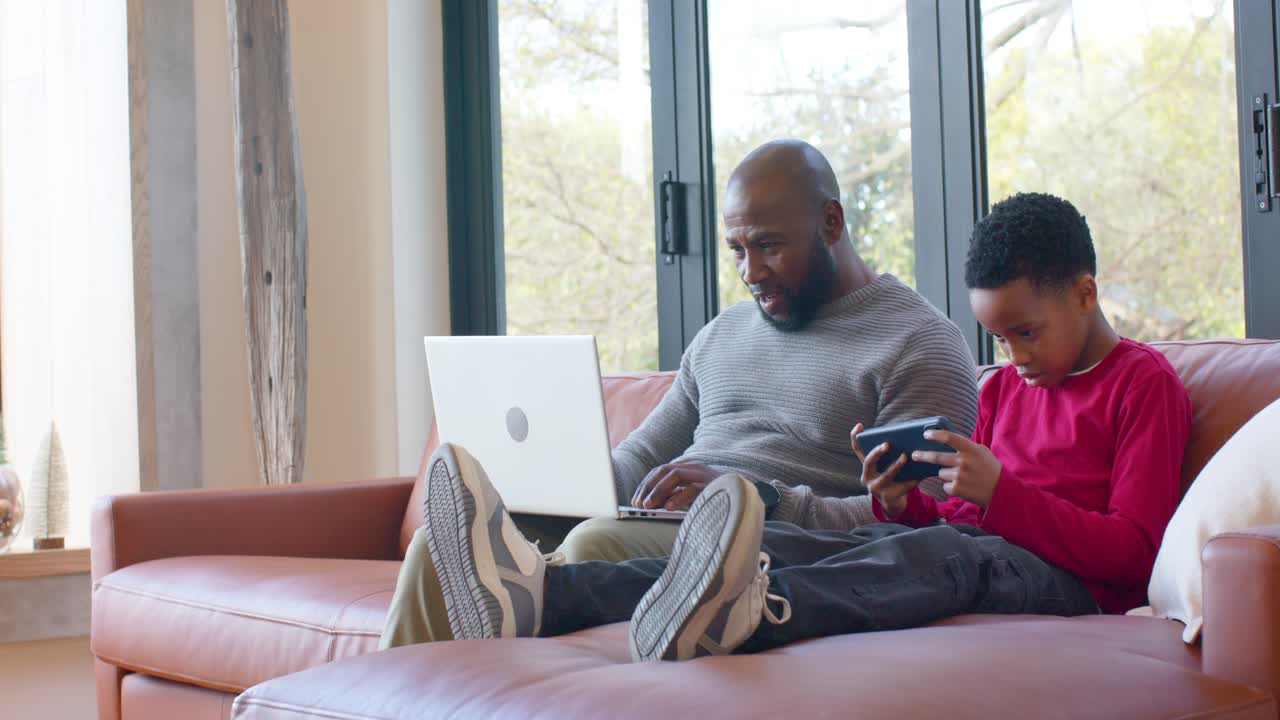 African American father glancing at son tapping device, lifting laptop and sharing screen on sofa