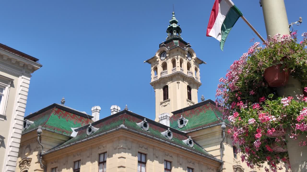 Hungary’s Szeged City Hall exterior shot featuring a vibrant green roof with foreground flowers and a waving flag