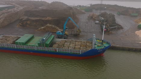 aerial establishing view of wood terminal crane loading timber into the cargo ship, port of liepaja , lumber log export, overcast day with fog and mist, drone shot moving forward, tilt down