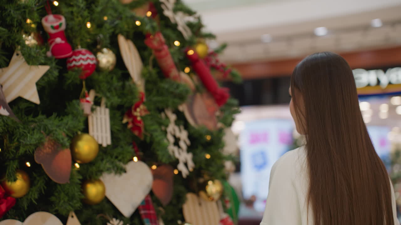 Young woman standing in front of Christmas tree, looking excited and amazed with bokeh light effect in background, shoppers walking by and building inscription visible in vibrant mall