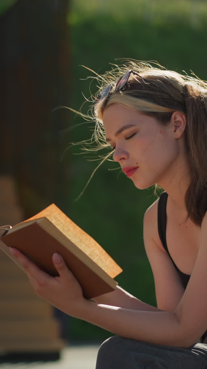 una dama sentada al aire libre lee un libro mientras el viento le sopla el cabello y levanta las páginas del libro, con la luz del sol reflejándose en ella, el fondo borroso presenta una escalera y vegetación