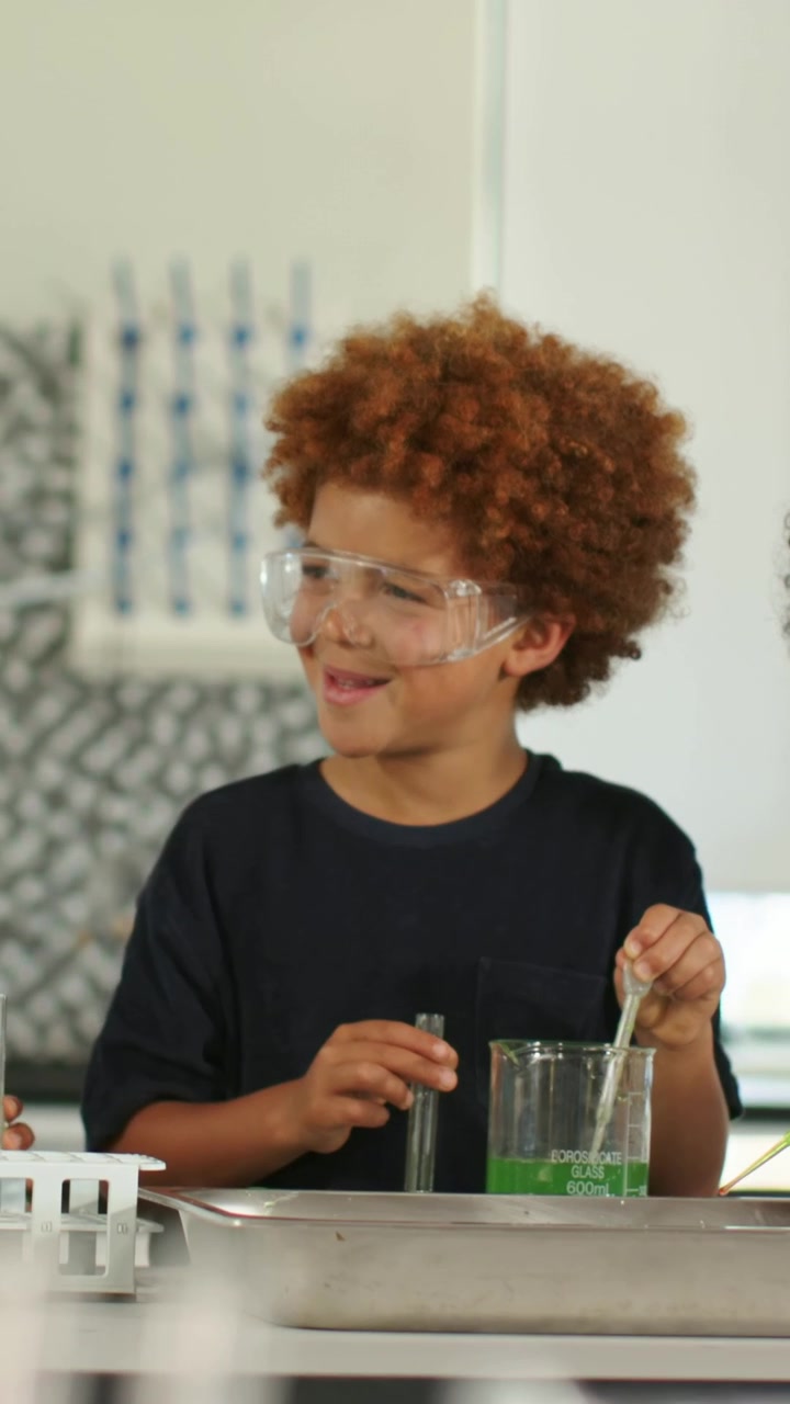 Vertical video: Stirring boy in shirt with goggles at lab with green beaker and test-tube for class