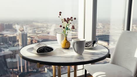 a small table set with flowers coffee cups and plates next to large windows in a downtown high rise condo