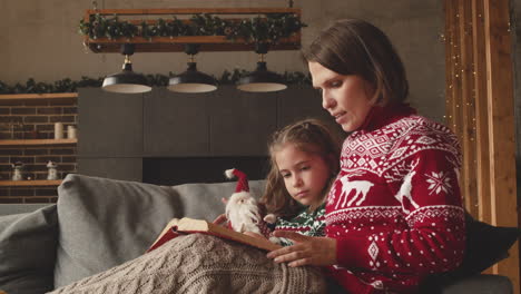 lovely mother sitting on couch with adorable daughter and reading her fairy tale on christmas day at home 2