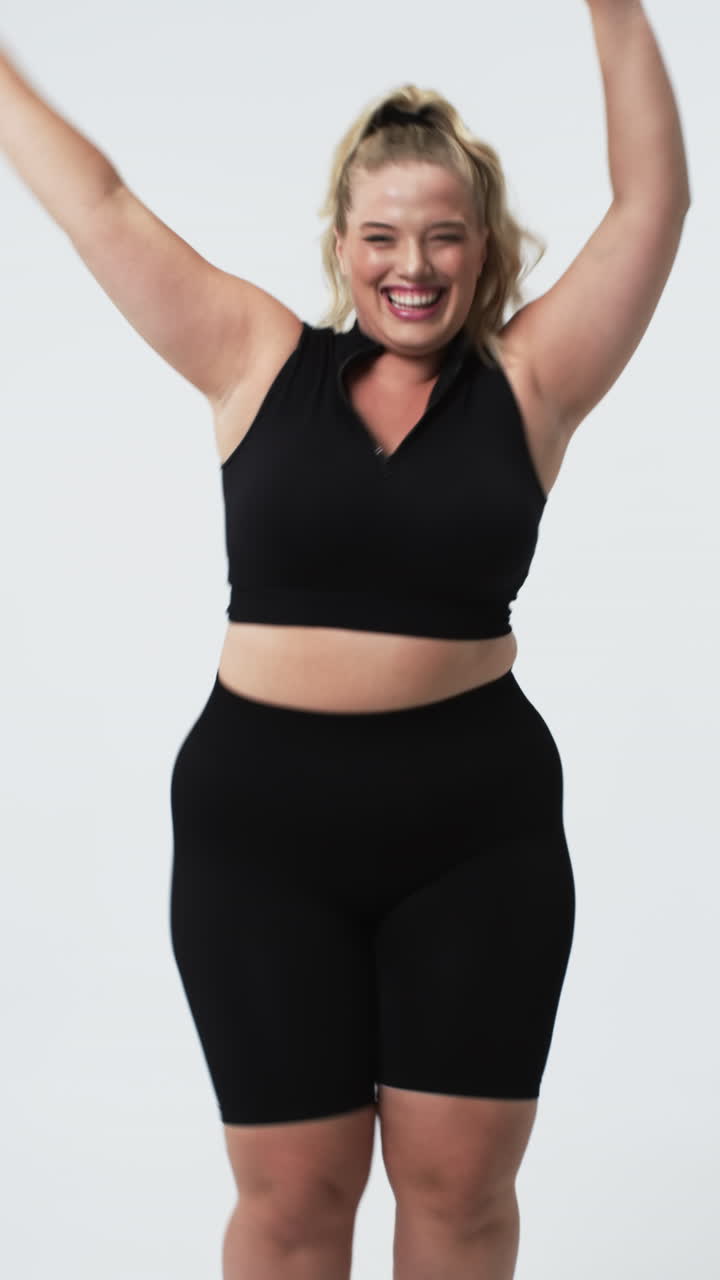 Vertical video: Smiling plus size woman in activewear celebrating body positivity with arms raised