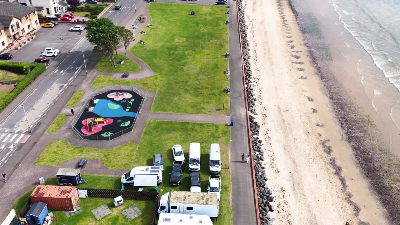 Drone flies above seaside park, playground, and caravans beside beach under bright daylight