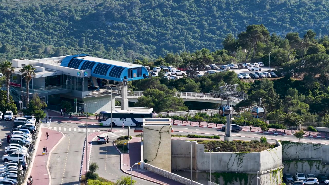 Rekaman Drone yang Menampilkan Bangunan Terminal Atas Modern Sistem Kereta Gantung Umum di Gunung Karmel, Dikelilingi oleh Jalan, Tempat Parkir, dan Hutan Hijau
