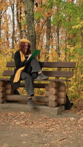 artista femenina dibujando sentada en un banco de madera del parque con una bolsa en el suelo en consuelo rodeada de árboles dorados de otoño en un entorno al aire libre pacífico, inmersa en la creatividad, con boina amarilla y bufanda