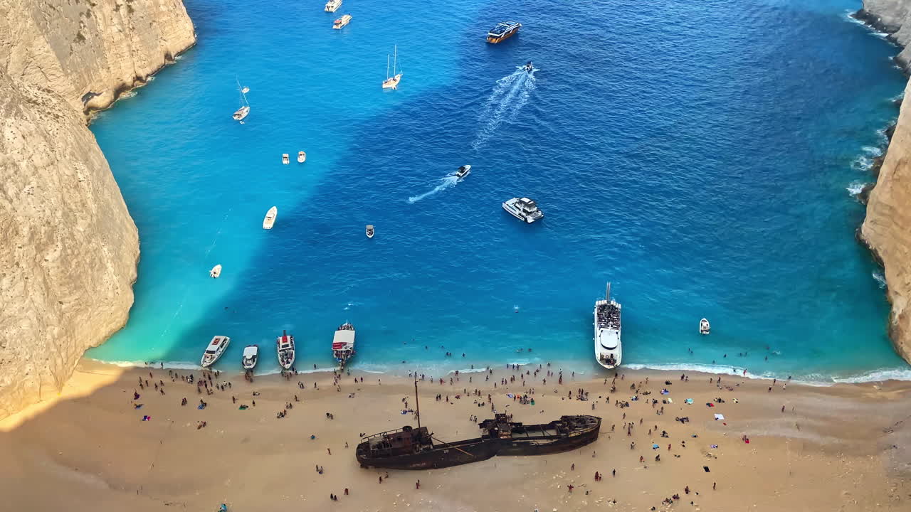Aerial drone view of the Navagio beach on the Ionian Sea coast of Zakynthos, Greece. Moored boats and resting people, rusty ship, blue water. Slow motion