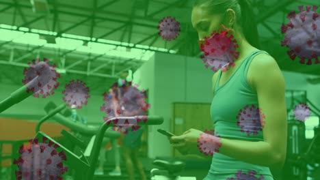 Covid-19-cells-floating-against-woman-wearing-face-mask-using-smartphone-at-the-gym