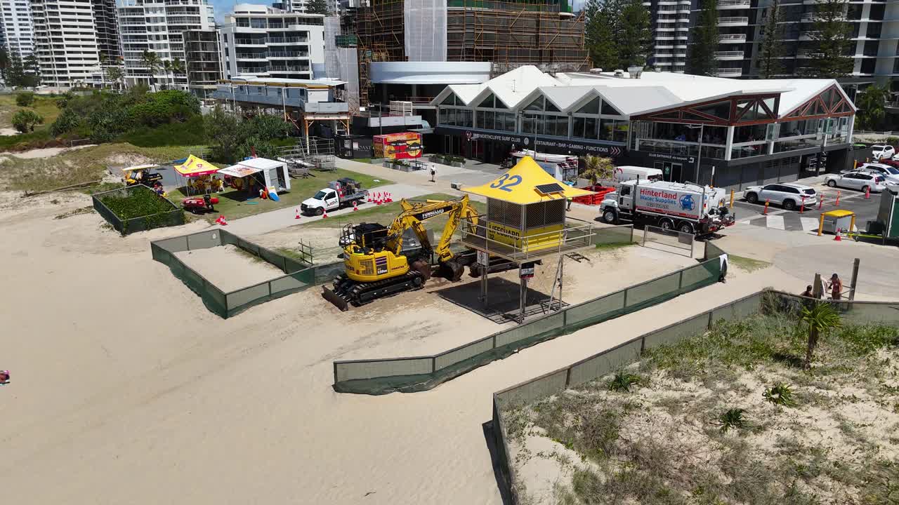 Aerial view of construction vehicles and life-saving tower on a sandy beach, surrounded by urban buildings