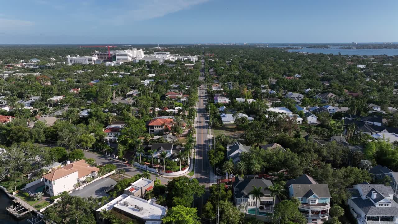Straight street in residential neighborhoods with lush greenery. The cityscape stretches under a clear blue sky, showcasing a blend of urban and natural beauty. Sarasota, Florida. Aerial wide shot.
