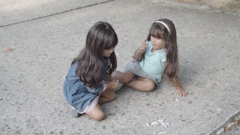 dos niñas latinas sentadas en el asfalto, dibujando con tizas y hablando