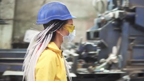 Woman-worker-in-yellow-and-transparent-protecting-glasses,-hard-hat-and-mask-inspect-equipment-on-recycle-plant.-Press-machine-with-used-plastic-bottles-and-differen-plastic-garbage.-Footage-of-automized-process