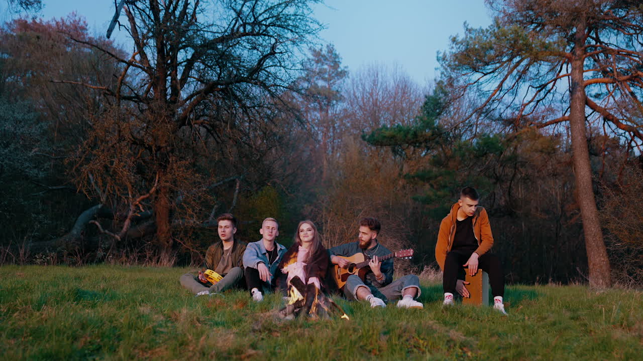 Happy friends relax in nature. Young people sitting by the campfire and singing songs. Guitarist is playing the guitar. Friendship concept.