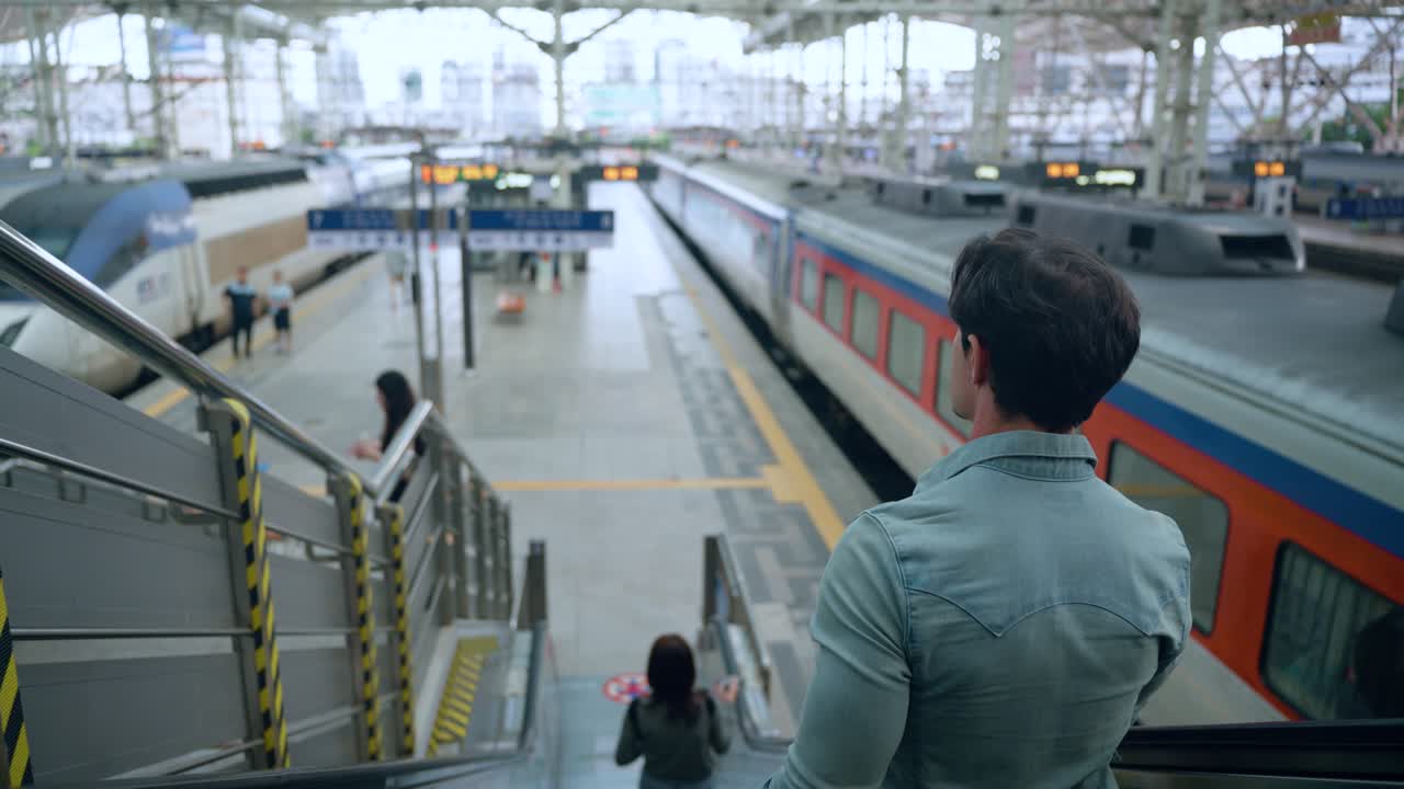 Australian Man Traveler Riding Escalator Down to Platforms of Seoul Train Station, View Of Railway Tracks and Locomotives - over shoulder view