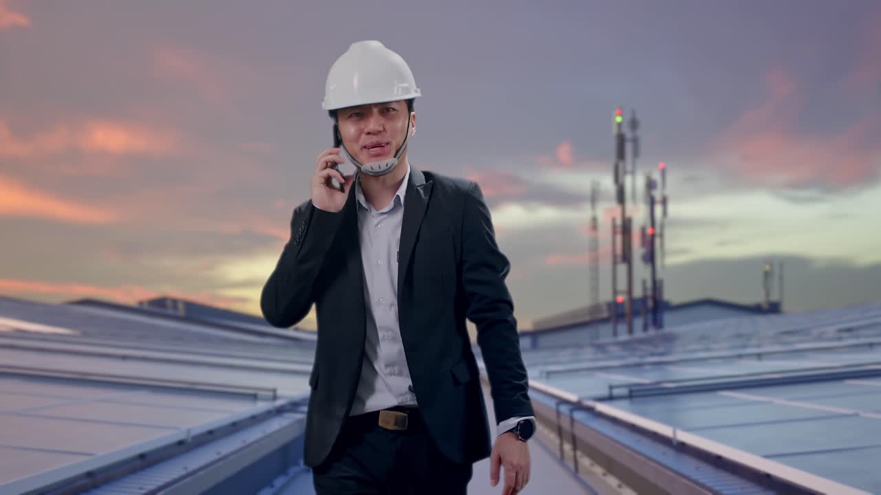 Vista frontal de empresario asiático hablando por smartphone mientras camina entre paneles solares industriales en la azotea