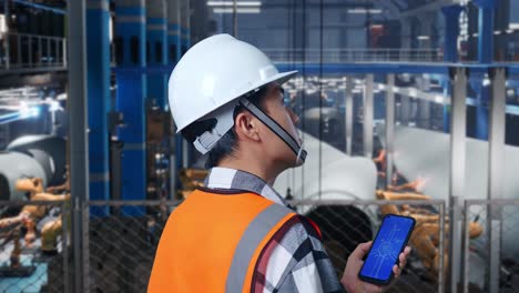 vista cercana de atrás de un ingeniero masculino asiático con casco de seguridad trabajando en un teléfono inteligente con el plan de la turbina eólica en una fábrica de turbinas eólicas mientras el brazo robótico trabaja