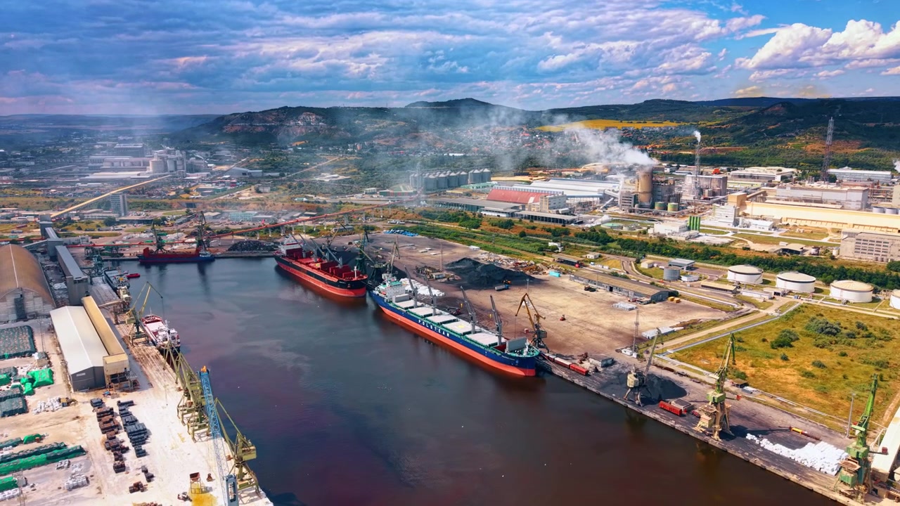 Varna, Bulgaria, 16 de junio de 2026: Graneleros atracados frente a obras industriales. Vista oblicua de dron que muestra buques de carga rojos, pilas de carbón, grúas y edificios de fábricas humeantes junto al agua.