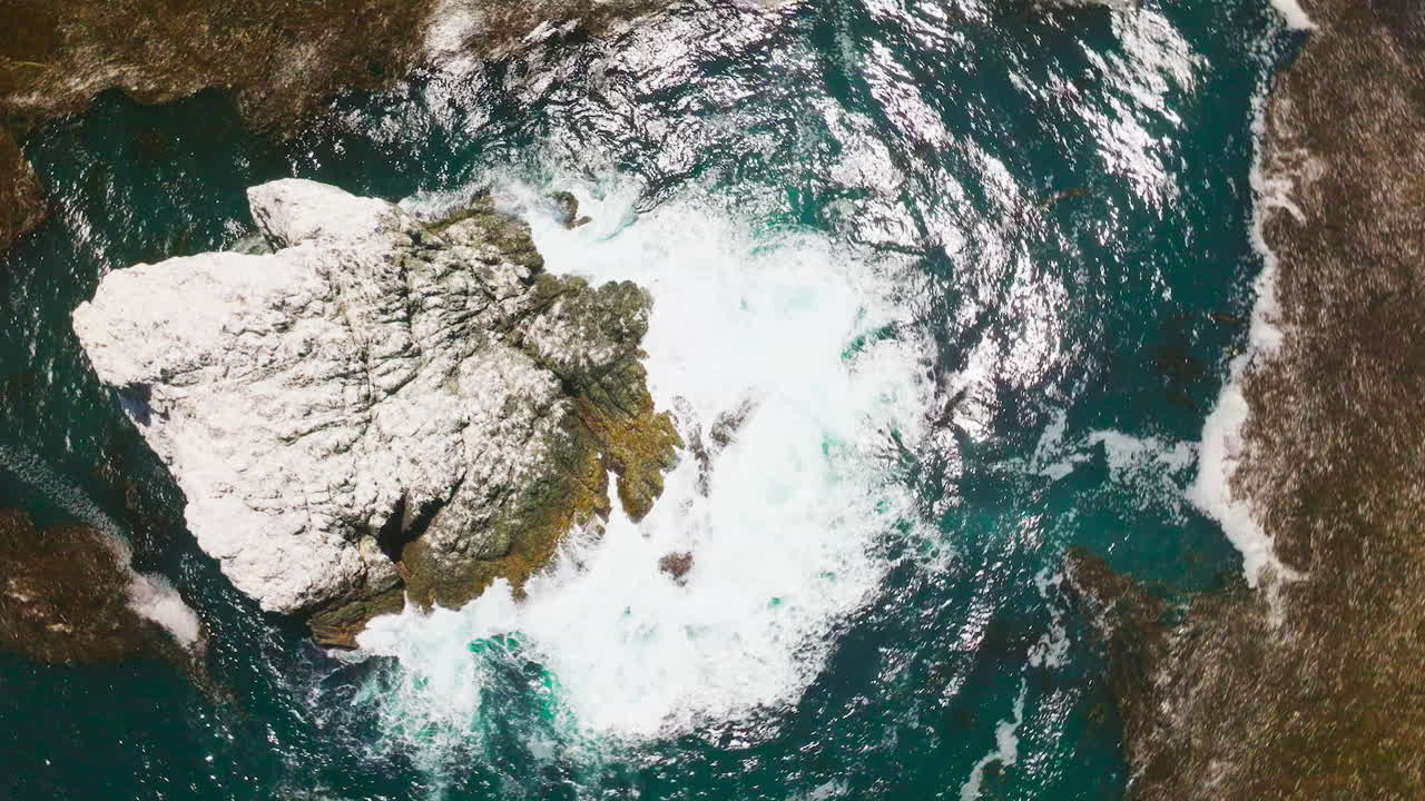 Craggy ragged rock in the blue ocean. White waves splashing by the cliff covered with sea salt. Top view.