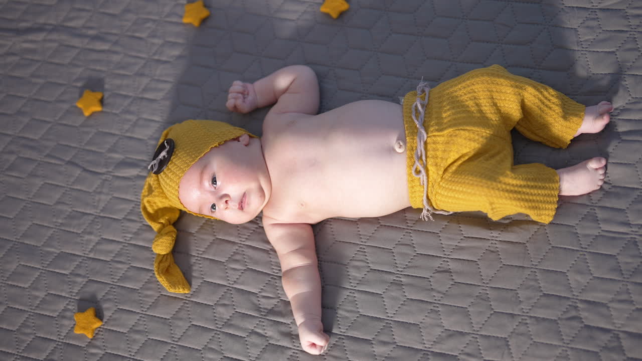 Kid in yellow hat with big button and wide pants is on the bed. Toddler in a funny suit with naked belly resting on a big bed. View from above.