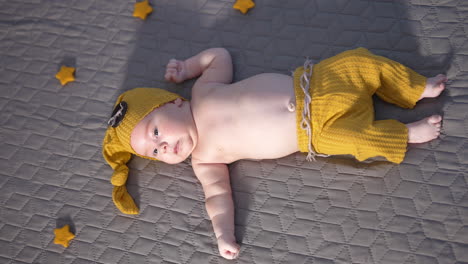 kid in yellow hat with big button and wide pants is on the bed. toddler in a funny suit with naked belly resting on a big bed. view from above.