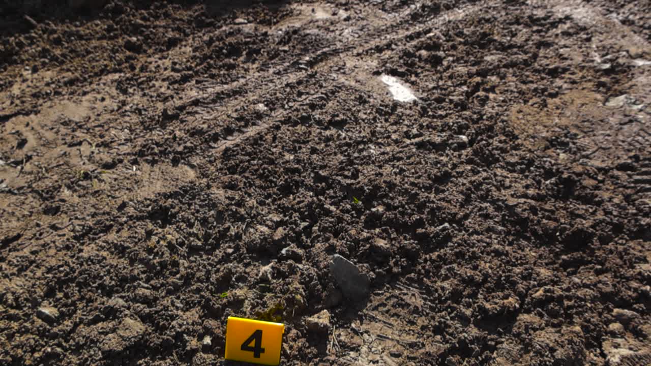 Top down view of a boot or shoe footprint as forensic evidence next to yellow colored police crime scene marker during sunny day time in muddy forest countryside road with tiretracks visible also