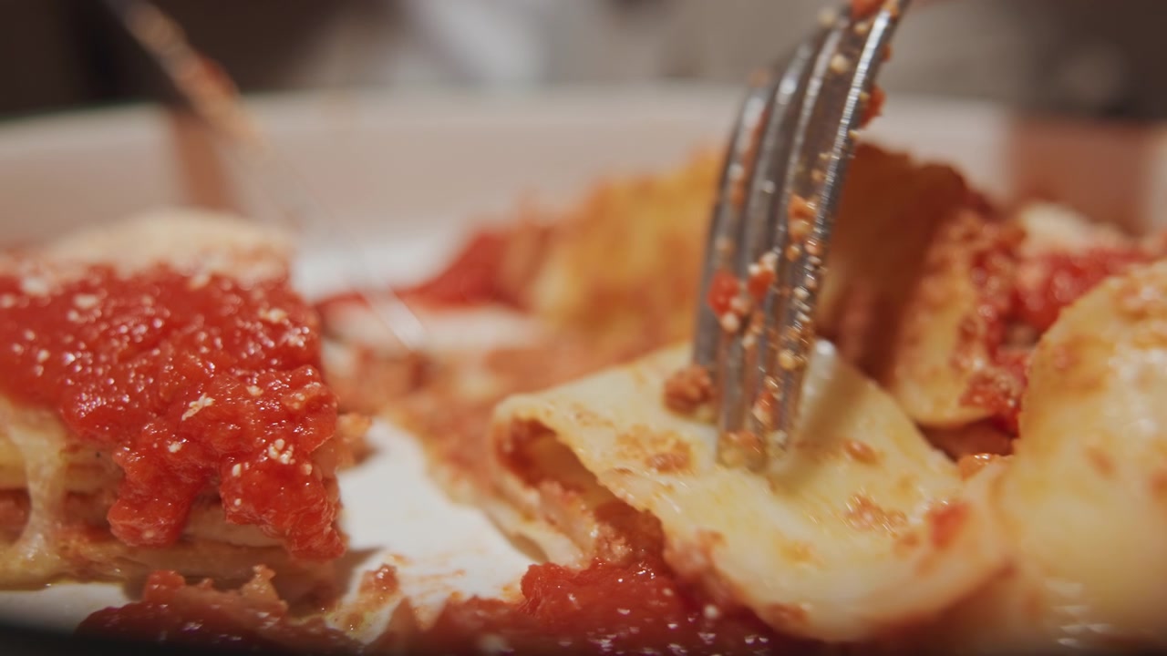 Primo piano di lasagne tagliate con forchetta e coltello che rivelano strati di salsa al formaggio, ripieno di carne e condimento al pomodoro. La forchetta solleva le fette dal piatto mostrando i dettagli della consistenza morbida. Ambiente interno.