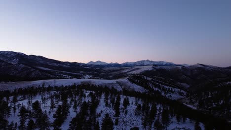 Flying-over-pine-trees-in-winter-towards-the-Rocky-Mountains-during-blue-hour,-aerial