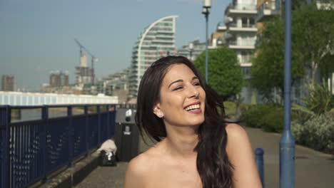 Medium-shot-of-an-attractive-and-playful-latina-woman-with-black-wavy-hair-walking-on-the-banks-of-the-Thames-river-in-London,-looking-at-the-camera,-spinning,-happy-with-a-beautiful-smile