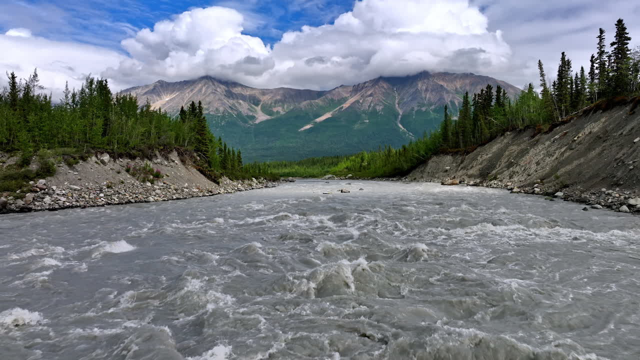 Turbulent mountain river with grey dirty water. Rocks with fluffy white cloudscape on top at backdrop. Alaska wilderness