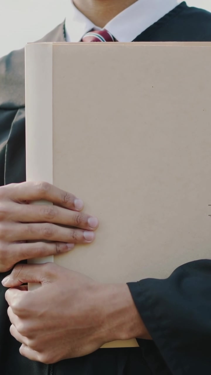 Wideo pionowe: Pozujący absolwent przesuwający dłonie po beżowej teczce na zewnątrz, ubrany w togę, białą koszulę i krawat