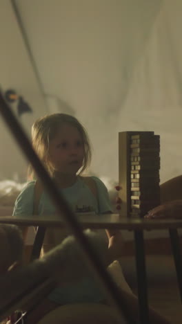 cheerful mother prepares wooden tower for playing jenga with son and daughter. family sitting at small table in cozy tourist chairs inside glamping camp in nature