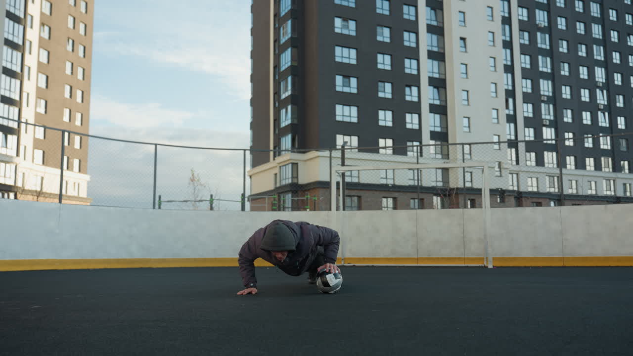 atleta realizando flexiones en una arena deportiva al aire libre con colocación alterna de la mano en la pelota de fútbol, mostrando coordinación, con postes de gol y edificios residenciales en el fondo urbano