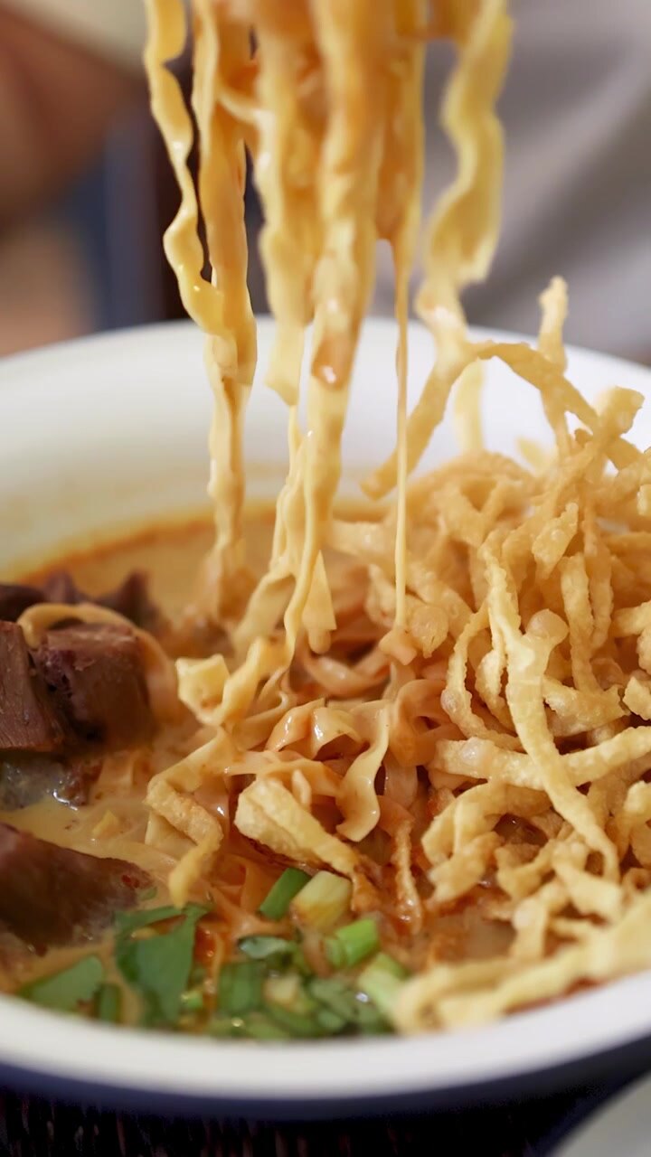 Una mano usa le bacchette per sollevare spaghetti all'uovo da una ciotola di zuppa di noodle al curry di manzo thailandese del nord, con illuminazione vibrante e profondità di campo ridotta