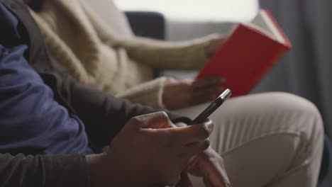 close up of couple at home sitting on sofa in lounge with woman reading book and man looking at mobile phone