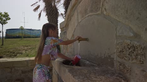 young child washing her red drinking cup under a water spout outside