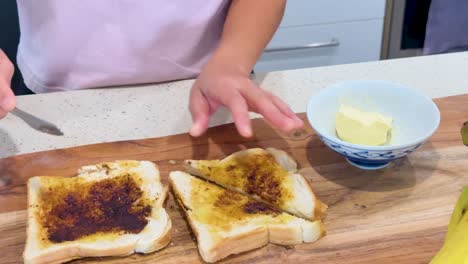 hands slice white bread toast topped with dark savory spread and butter on a wooden board in a brightly lit modern kitchen