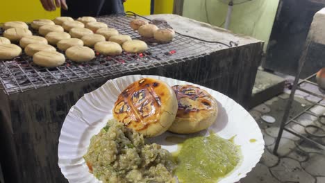 in the backdrop, a vendor is seen baking the litti in a hot iron grill while serving it with baigan bharta and green in a paper plate