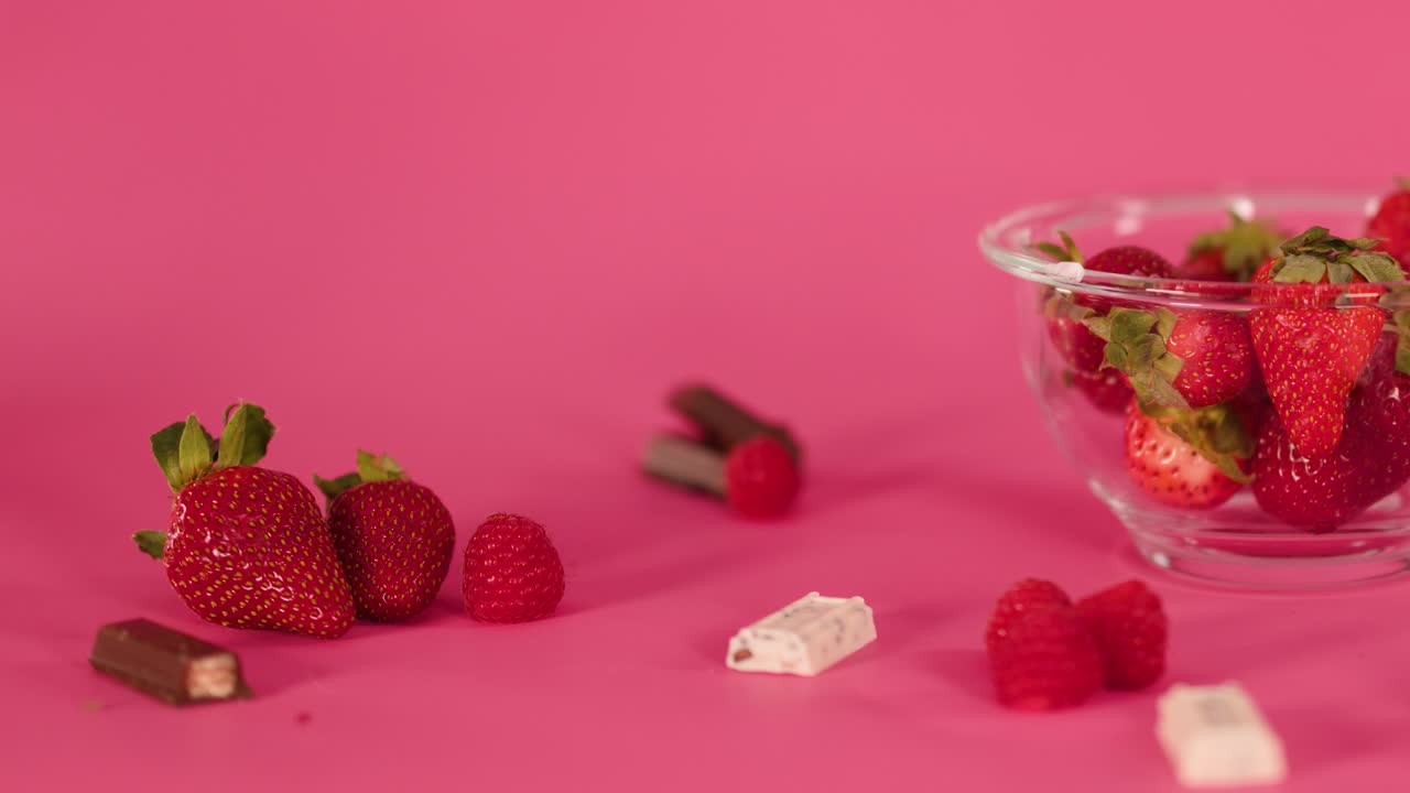 Hand arranges strawberries, raspberries, and assorted candies on vibrant pink surface with soft lighting