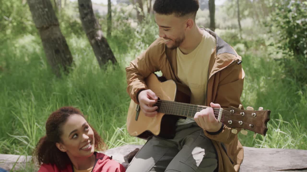 feliz pareja afroamericana sentada en el tronco de un árbol y tocando la guitarra en el bosque, cámara lenta