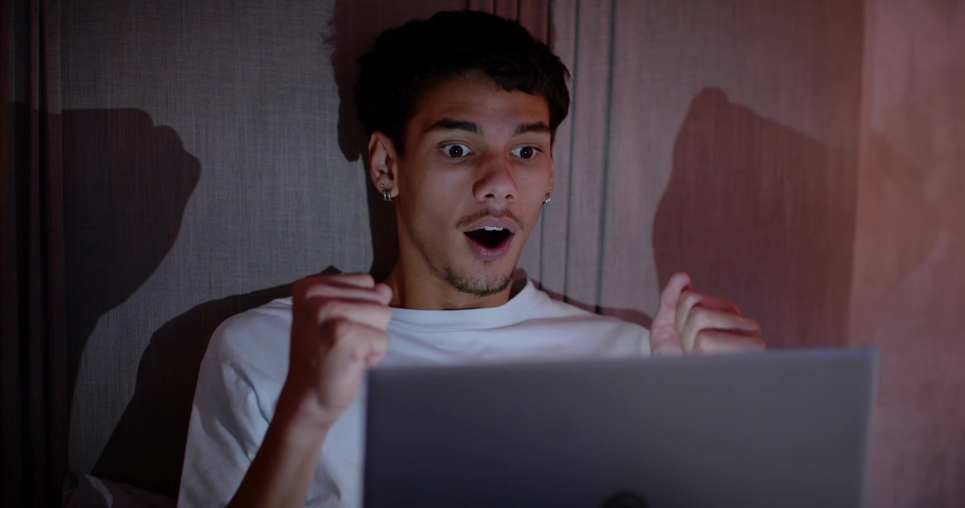 African American man in twenties on bed with laptop, lifting head, reacting to surprising message